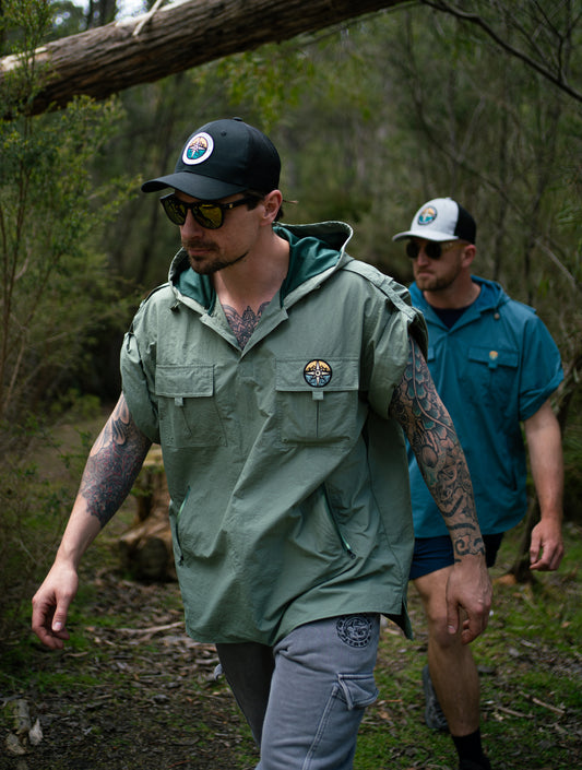 Two men walking in a forest, one wearing a green jacket and cap with a logo.